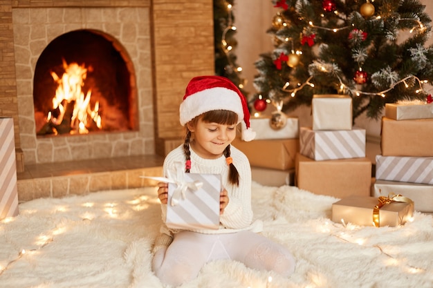 Glimlachend meisje met witte trui en kerstman hoed, zittend op de vloer in de buurt van kerstboom, geschenkdozen en open haard, cadeau van ouders in handen houdend.