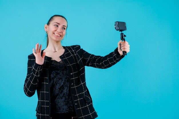 Glimlachend meisje neemt selfie met minicamera door hallo gebaar te tonen op blauwe achtergrond