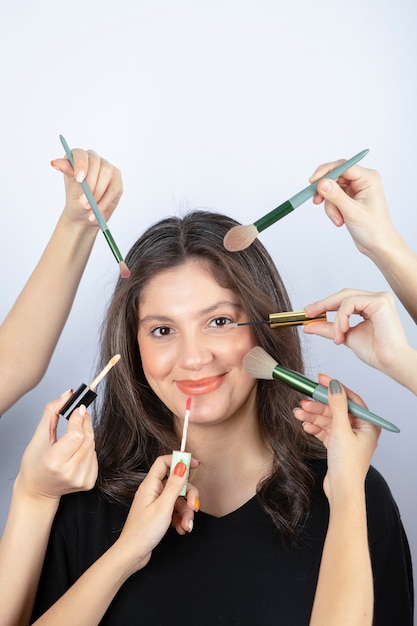 Glimlachend meisje omringd door handen van make-upartiesten met borstels, lippenstift en mascara in de buurt van haar gezicht.