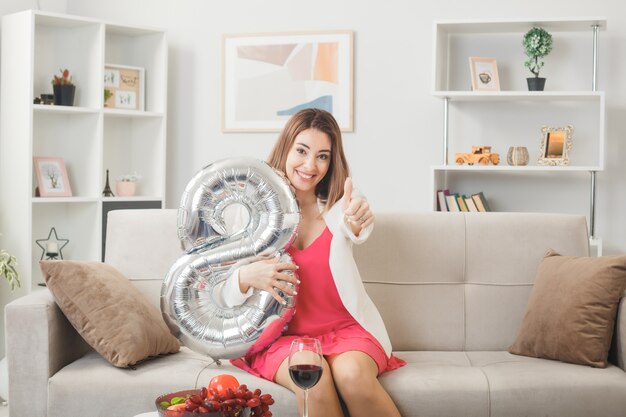 Glimlachend met duim omhoog vrouw op gelukkige vrouwendag met nummer acht ballon zittend op de bank in de woonkamer