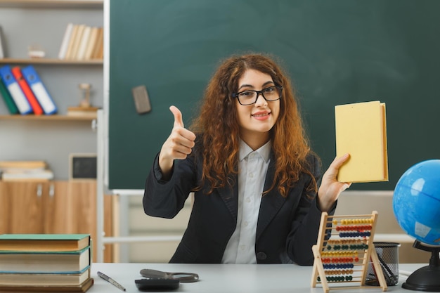 Glimlachend met duimen omhoog jonge vrouwelijke leraar die een bril draagt en een boek vasthoudt dat aan een bureau zit met schoolhulpmiddelen in de klas