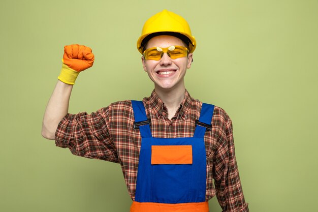 Glimlachend met gesloten ogen die de hand opheft, jonge mannelijke bouwer met uniform en handschoenen met bril