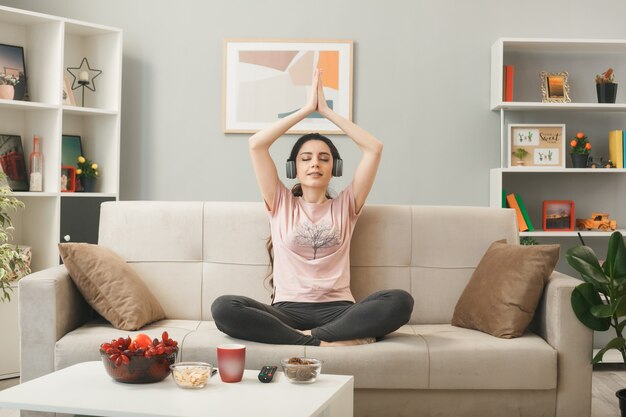 Glimlachend met gesloten ogen jong meisje met een koptelefoon op die yoga doet, zittend op de bank achter de salontafel in de woonkamer