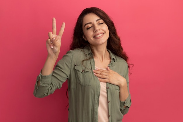 Glimlachend met gesloten ogen jong mooi meisje dat olijfgroen t-shirt draagt dat vredesgebaar toont dat op roze muur wordt geïsoleerd