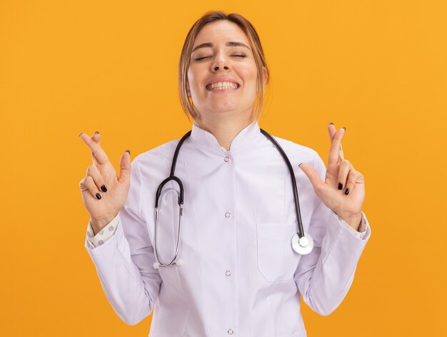 Glimlachend met gesloten ogen jonge vrouwelijke arts die medische mantel draagt met een stethoscoop die vingers kruist die op gele muur worden geïsoleerd