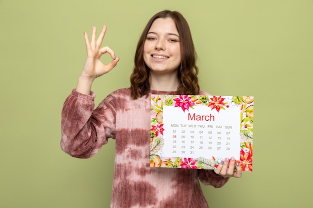 Glimlachend met goed gebaar mooi jong meisje op gelukkige vrouwendag met kalender geïsoleerd op olijfgroene muur