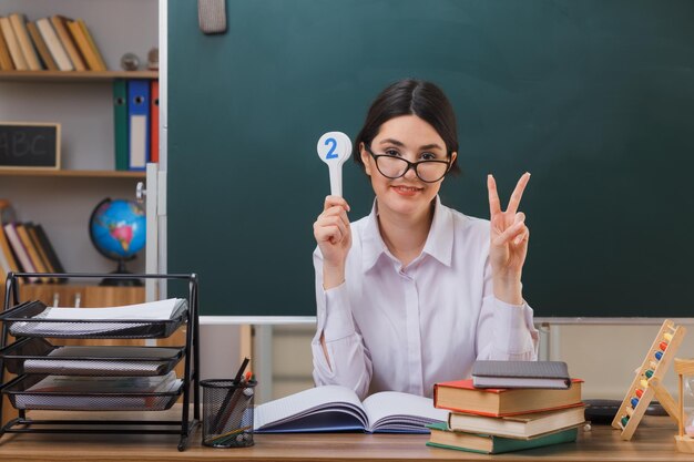 glimlachend met vredesgebaar jonge vrouwelijke leraar die een bril draagt met nummer plezier zittend aan een bureau met schoolhulpmiddelen in de klas