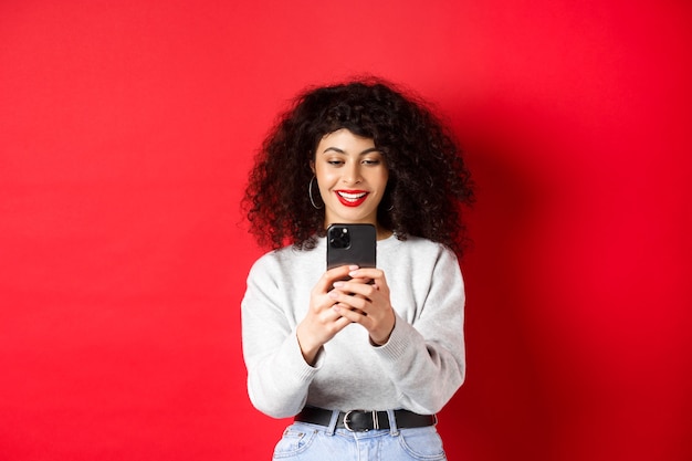 Glimlachend modern meisje dat foto's maakt op een smartphone, naar het scherm kijkt en video opneemt, staande tegen een rode achtergrond