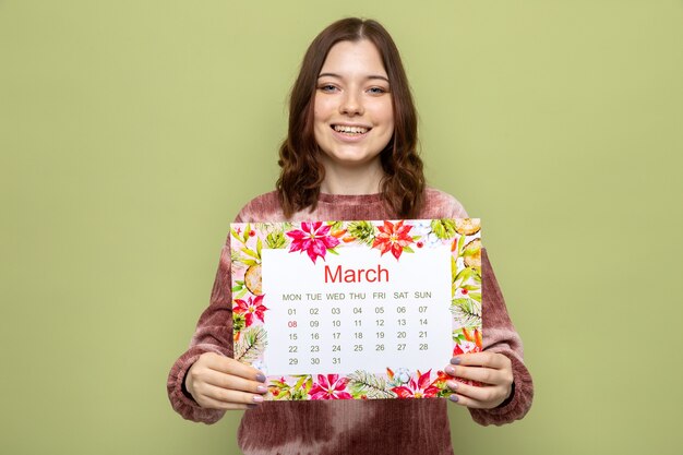 Glimlachend mooi jong meisje op een gelukkige vrouwendag die kalender uithoudt naar camera geïsoleerd op olijfgroene muur