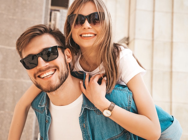 Glimlachend mooi meisje en haar knappe vriendje in casual zomer kleding. Man die zijn vriendin op de rug draagt en zij die haar handen opheft.