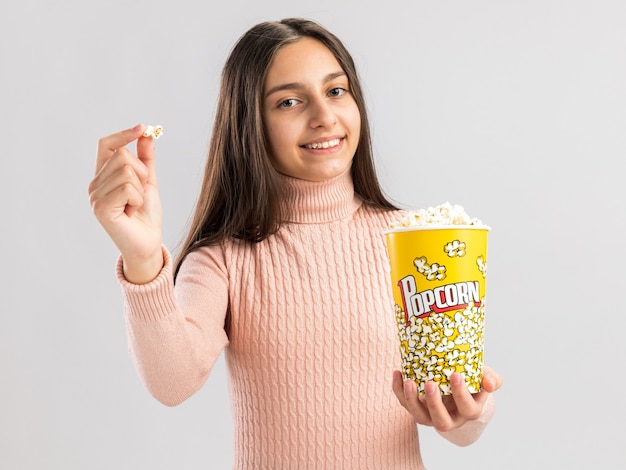 Glimlachend mooi tienermeisje dat naar de voorkant kijkt en een emmer popcorn en popcornstuk uitrekt naar camera geïsoleerd op een witte muur