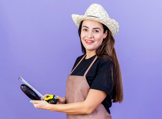 Glimlachend mooi tuinman meisje in uniform dragen tuinieren hoed aubergine meten met meetlint geïsoleerd op blauw