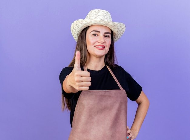 Glimlachend mooi tuinman meisje in uniform dragen tuinieren hoed duim opdagen en hand zetten heup geïsoleerd op blauwe achtergrond