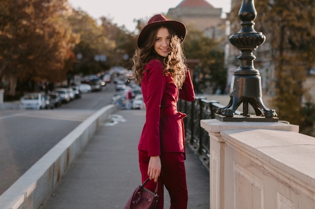 Glimlachend mooie stijlvolle vrouw in paars pak wandelen in de stad straat, lente zomer herfst seizoen modetrend dragen hoed, portemonnee te houden