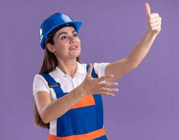 Glimlachend op zoek naar jonge bouwersvrouw in uniform met duim omhoog geïsoleerd op paarse muur