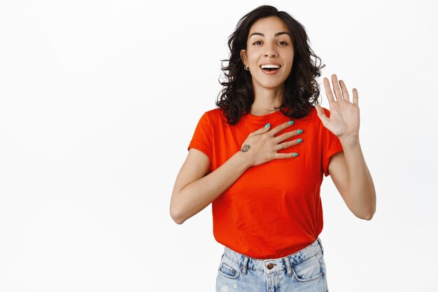 Glimlachend opgewonden meisje dat vrijwilligerswerk doet, zeg dat ik ben, hand opsteken en zichzelf voorstellen, de waarheid vertellen of belofte doen, staande op een witte achtergrond