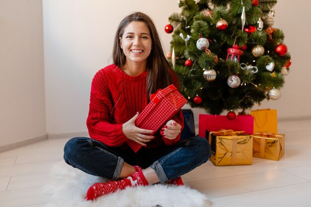 Glimlachend opgewonden mooie vrouw in rode trui om thuis te zitten bij de kerstboom uitpakken van cadeautjes en geschenkdozen