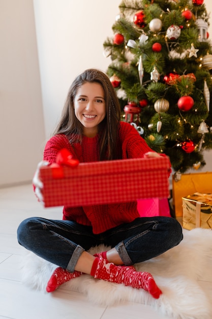 Glimlachend opgewonden mooie vrouw in rode trui om thuis te zitten bij de kerstboom uitpakken van cadeautjes en geschenkdozen