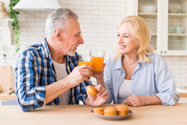 Glimlachend portret van een hoger paar die de glazen sap en muffins op lijst in de keuken roosteren