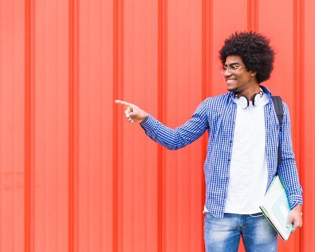 Glimlachend portret van een jonge mannelijke student die vinger richt op iets die zich tegen rode muur bevinden