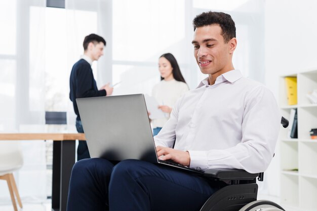 Glimlachend portret van een jonge zakenmanzitting op rolstoel die laptop met zijn collega met behulp van bij achtergrond