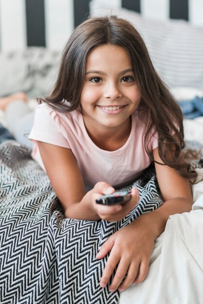 Glimlachend portret van meisje die op bed liggen die afstandsbediening met behulp van