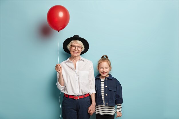 Glimlachend roodharig meisje komt oma feliciteren met Moederdag, draagt een gestreepte trui en een spijkerjasje. Blij dat senior dame in stijlvolle zwarte hoed, ballon draagt, hand houdt van kleine kleindochter