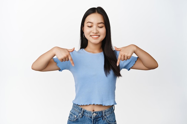 Glimlachend schattig Aziatisch meisje wijzend en naar beneden kijkend met reclame voor het lezen van promotekst onder de witte achtergrond