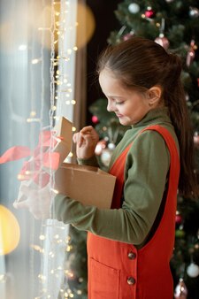 Glimlachend schattig brunette meisje in rode bodysuit permanent bij raam met verlichting en kijken naar kerst g...