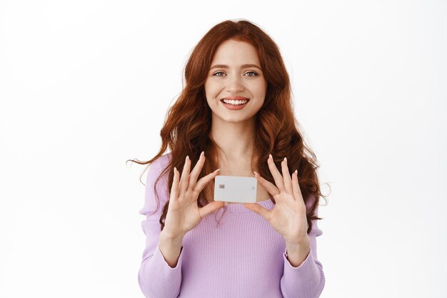 Gratis foto glimlachend schattig meisje met gemberhaar toont haar nieuwe bankcreditcard, contactloos winkelen in winkels, dingen kopen met aanbetaling, staande op een witte achtergrond