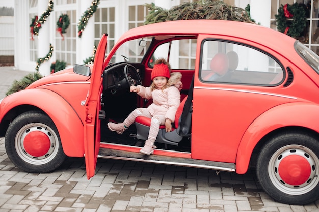 Glimlachend schattig wintermeisje in een rode hoed die in de auto zit en plezier heeft op middellange afstand fijne mooie vrouwelijke baby in warme kleding met positieve emotie buiten omringd door sneeuwvlokken die genieten van de kindertijd