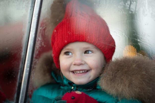 Glimlachend schattig wintermeisje in rode hoed die in de auto zit en plezier heeft op middellange afstand Fijne mooie vrouwelijke baby in warme kleding met positieve emotie buiten omringd door sneeuwvlokken die genieten van de kindertijd