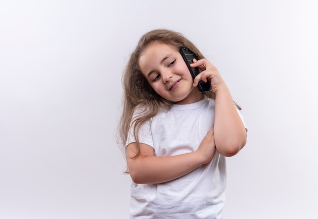Glimlachend schoolmeisje dat een wit t-shirt draagt, spreekt op telefoon op geïsoleerde witte achtergrond