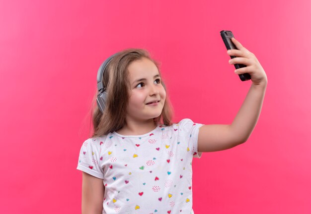 Glimlachend schoolmeisje die wit t-shirt in hoofdtelefoons dragen die selfie op geïsoleerde roze achtergrond nemen