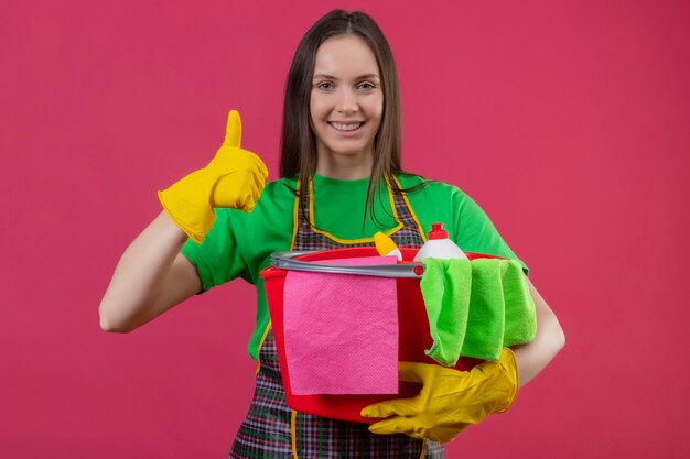 Glimlachend schoonmakend jong meisje die eenvormig in handschoenen dragen die schoonmakende hulpmiddelen haar duim op geïsoleerde roze achtergrond houden