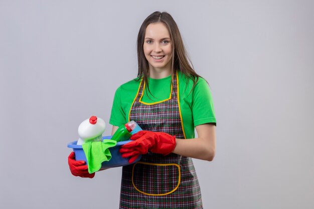 Glimlachend schoonmakend jong meisje die eenvormig in rode handschoenen dragen die schoonmakende hulpmiddelen op geïsoleerde witte achtergrond houden