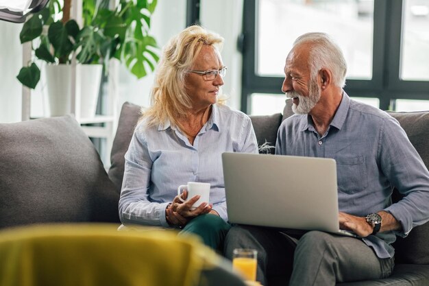 Glimlachend senior koppel dat computer gebruikt en met elkaar praat terwijl u thuis op de bank ontspant