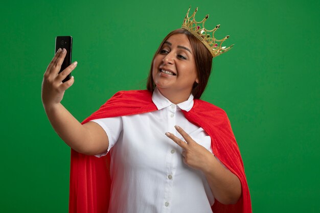 Glimlachend superheld vrouwtje van middelbare leeftijd dragen kroon nemen een selfie met vredesgebaar geïsoleerd op groene achtergrond