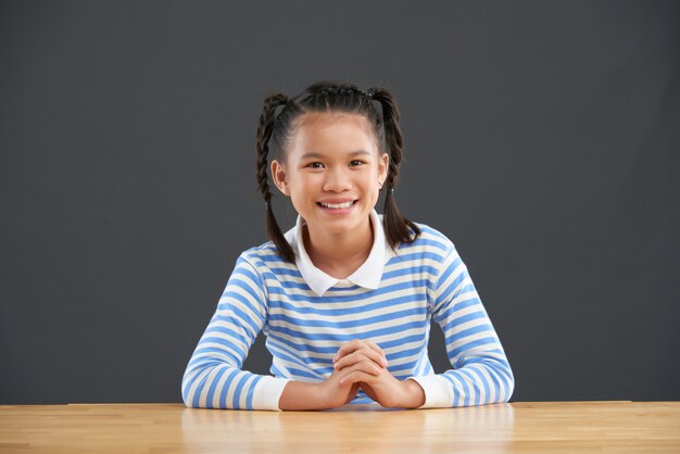 Glimlachend tiener Aziatisch schoolmeisje dat met vlechten bij bureau zit