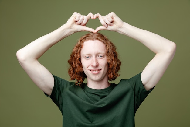 Glimlachend tonend hartgebaar jonge knappe kerel die groen t-shirt draagt dat op groene achtergrond wordt geïsoleerd