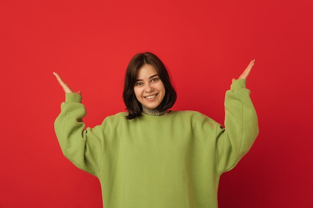 Glimlachend, verschijnt. portret van de blanke vrouw geïsoleerd op rode muur met copyspace. mooi vrouwelijk model in groene hoodie. concept van menselijke emoties, gezichtsuitdrukking