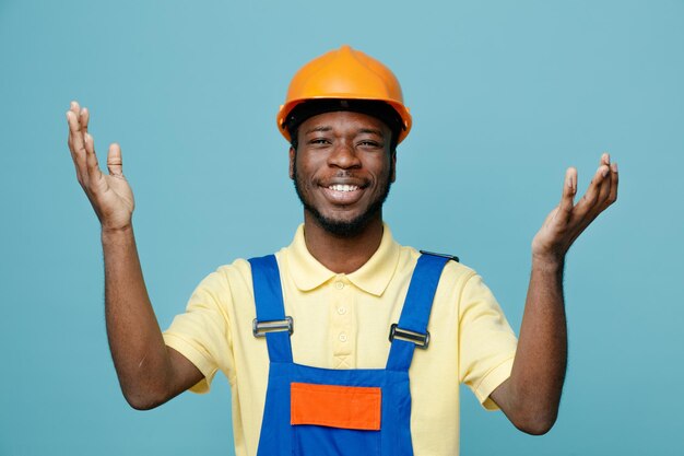 Glimlachend verspreidende handen jonge Afro-Amerikaanse bouwer in uniform geïsoleerd op blauwe achtergrond