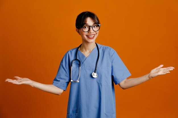 Glimlachend verspreidende handen jonge vrouwelijke arts dragen uniform fith stethoscoop geïsoleerd op oranje achtergrond