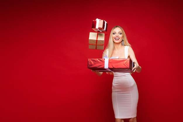 Glimlachend vrij jong meisje in feestelijke jurk en zwevende geschenkdozen op rode studio achtergrond met kopie ruimte voor advertentie. Hoge kwaliteit foto