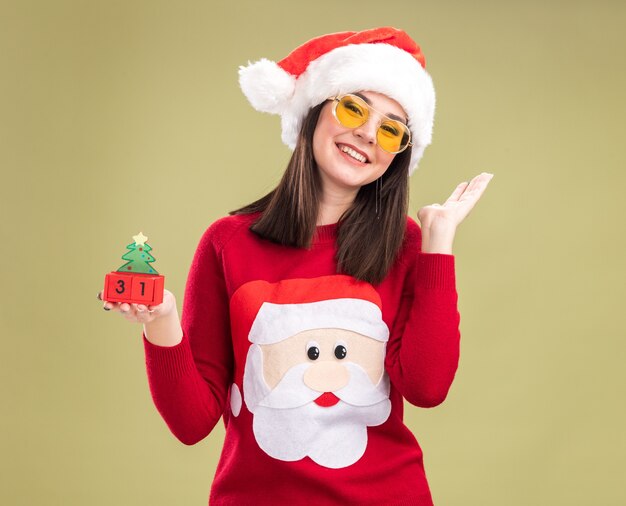 Glimlachend vrij Kaukasisch meisje dragen santa claus trui en hoofdband met bril kerstboom speelgoed met datum kijken camera weergegeven: lege hand geïsoleerd op olijfgroene achtergrond