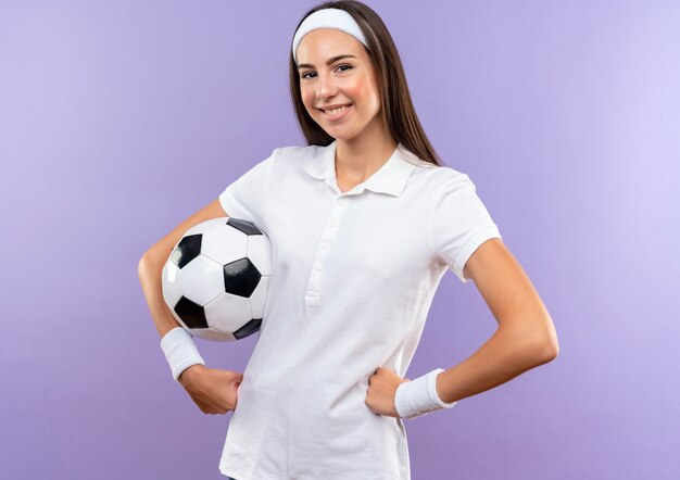 Glimlachend vrij sportief meisje dat hoofdband en polsbandje draagt met voetbal die handen op taille zet die op paarse ruimte wordt geïsoleerd