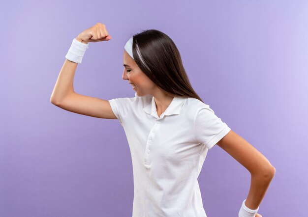 Glimlachend vrij sportief meisje die hoofdband en polsband dragen die zich in profielmening bevinden en sterk gebaren geïsoleerd op paarse ruimte