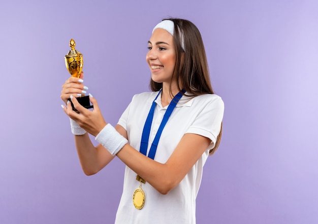 Glimlachend vrij sportief meisje met hoofdband en polsbandje en medaille houden en kijken naar beker geïsoleerd op paarse ruimte