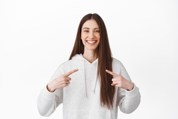 Glimlachend vrolijk brunette meisje wijst naar zichzelf, toont logo in het midden, perfecte witte glimlachtanden, zelfpromotie, staande tegen een witte muur