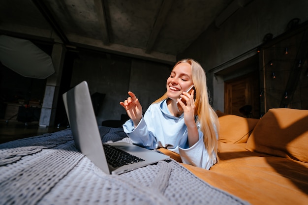 Glimlachende aantrekkelijke vrouw die op bed ligt dat door smartphone spreekt